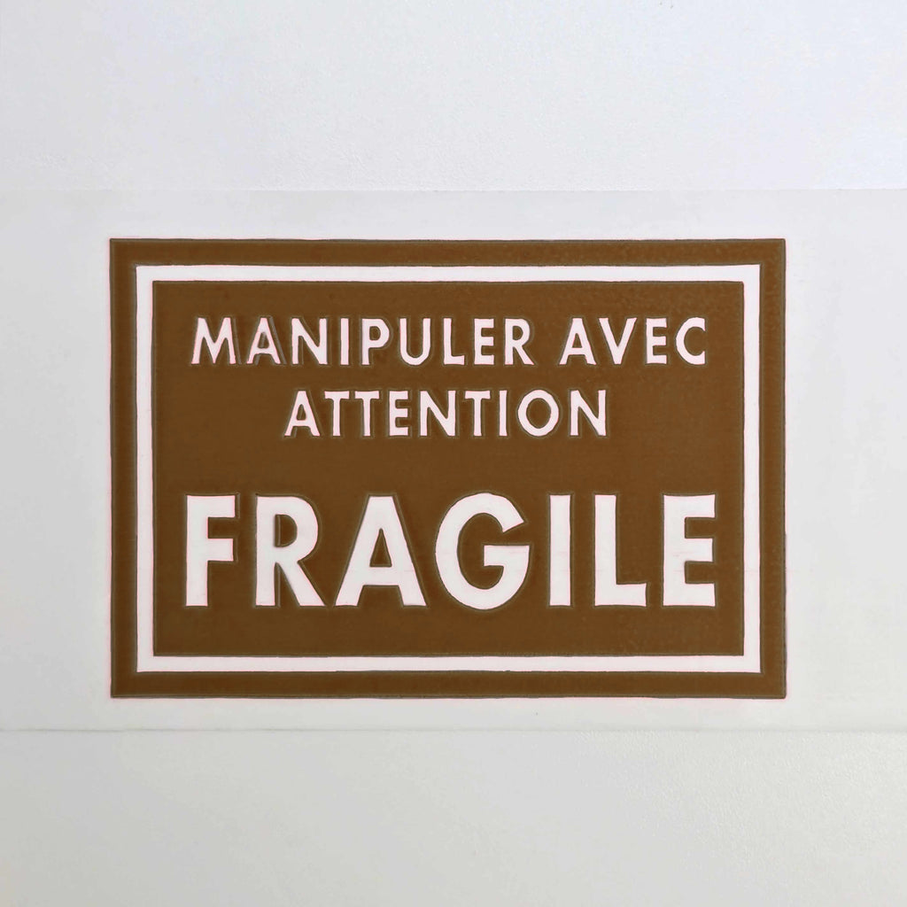 Brown sign with white text 'Manipuler Avec Attention Fragile' on a white background